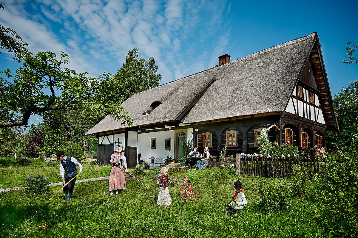 Zwei Erwachsene arbeiten in traditioneller Kleidung mit Sensen in einem Garten auf einer grünen Wiese. Drei Kinder in bunter Kleidung spielen daneben. Dahinter ist ein historisches Fachwerkhaus.  Auf der Veranda sitzen ein Mann und eine Frau und unterhalten sich. Der Himmel ist blau mit weißen Wolken.