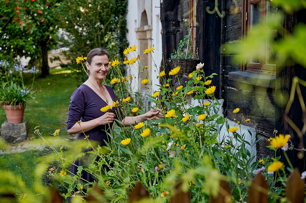 Eine Frau steht in einem Garten neben einem alten Fachwerkhaus. Sie lächelt und steht neben hohen gelben Blumen. Im Hintergrund sind grüne Bäume und eine Wiese. 