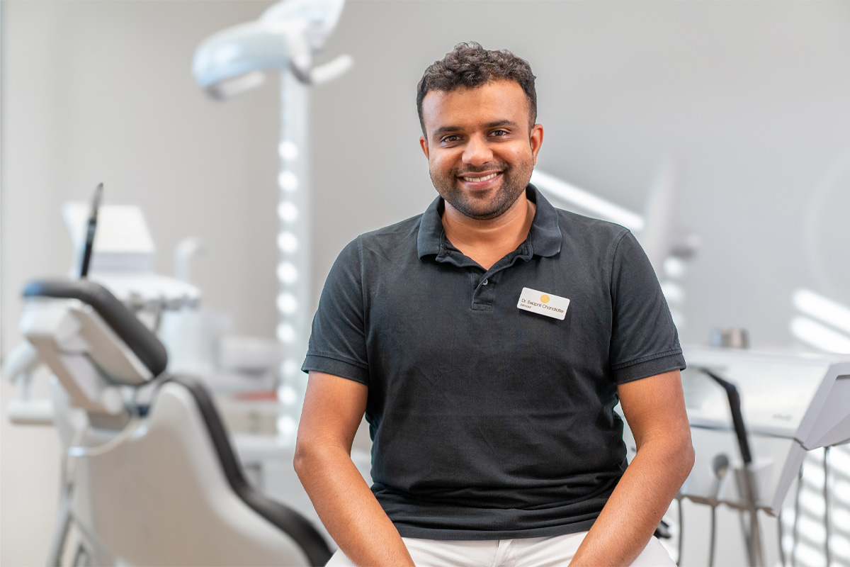 The image shows a man in a modern, bright treatment room. He is wearing a dark polo shirt with a name tag and is sitting smiling in front of a dentist&#39;s chair. The friendly atmosphere and professional setting suggest a dental practice.