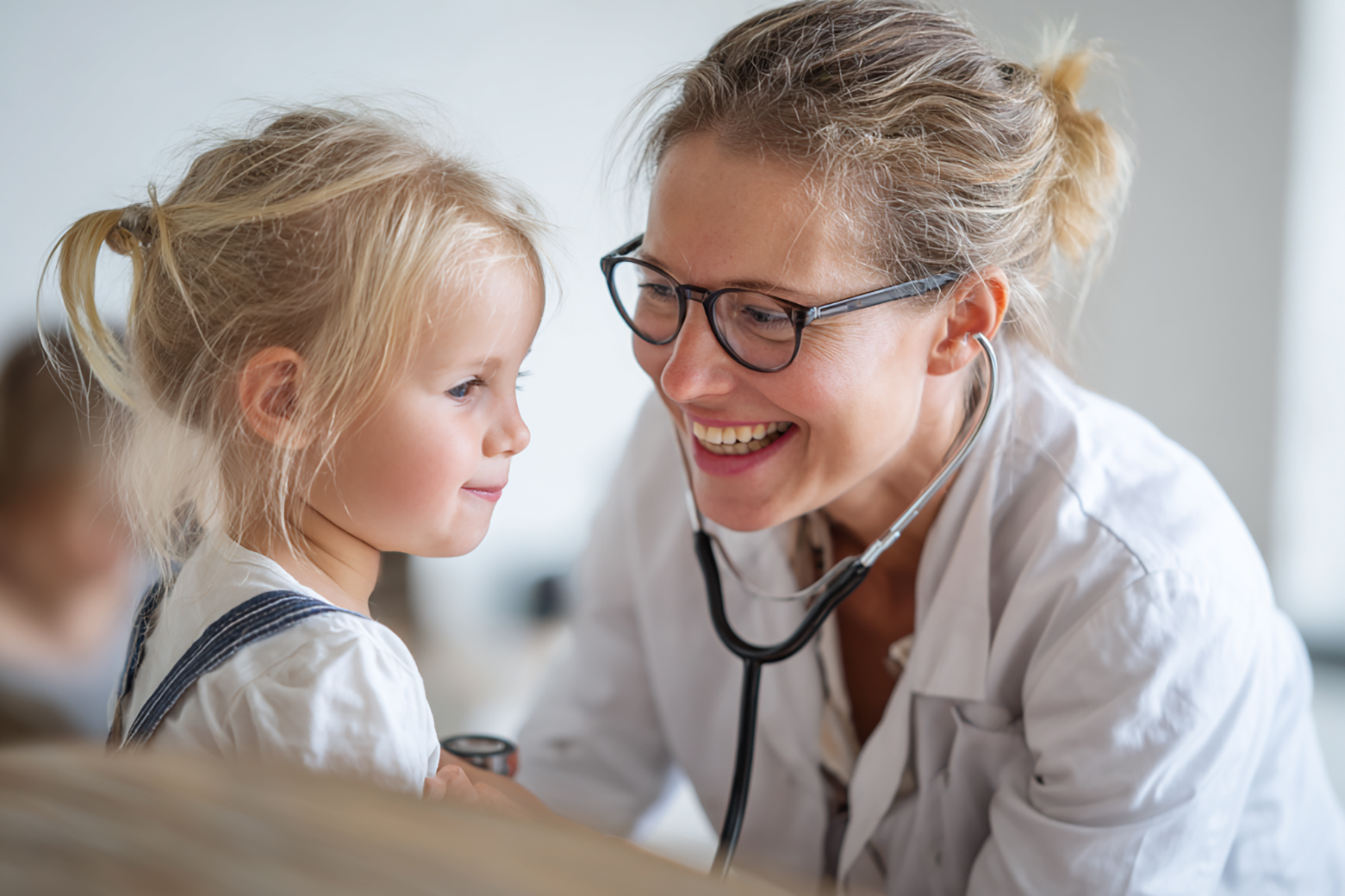 A smiling doctor stands in the foreground of a brightly lit practice or clinic. In the background, two medical professionals are discussing something on a tablet. The image conveys professionalism, trust, and teamwork in healthcare.