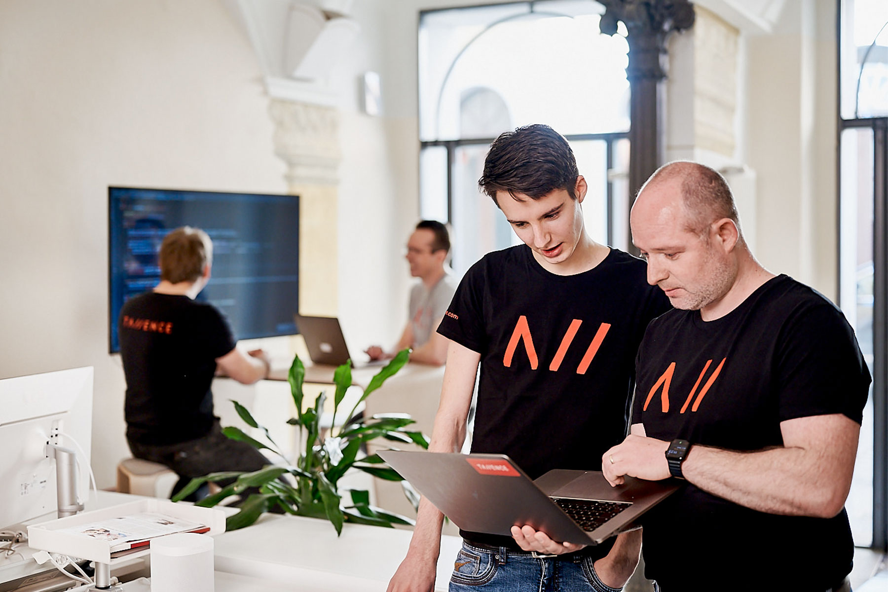 Zwei Männer in schwarzen T-Shirts mit rotem Logo stehen in einem modernen Büro und schauen gemeinsam auf einen Laptop. Im Hintergrund arbeiten zwei weitere Personen an Computern vor einem großen Bildschirm.