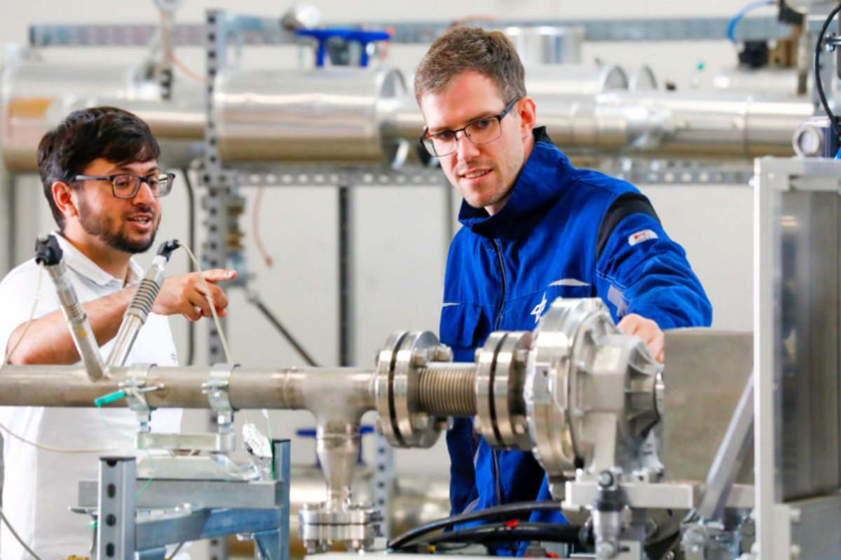 Zwei Männer stehen in einem Raum vor einer technischen Apparatur mit Rohren und Metallteilen. Einer trägt einen blauen Arbeitsanzug, der andere weiße Kleidung. Sie schauen beide auf die Apparatur. 