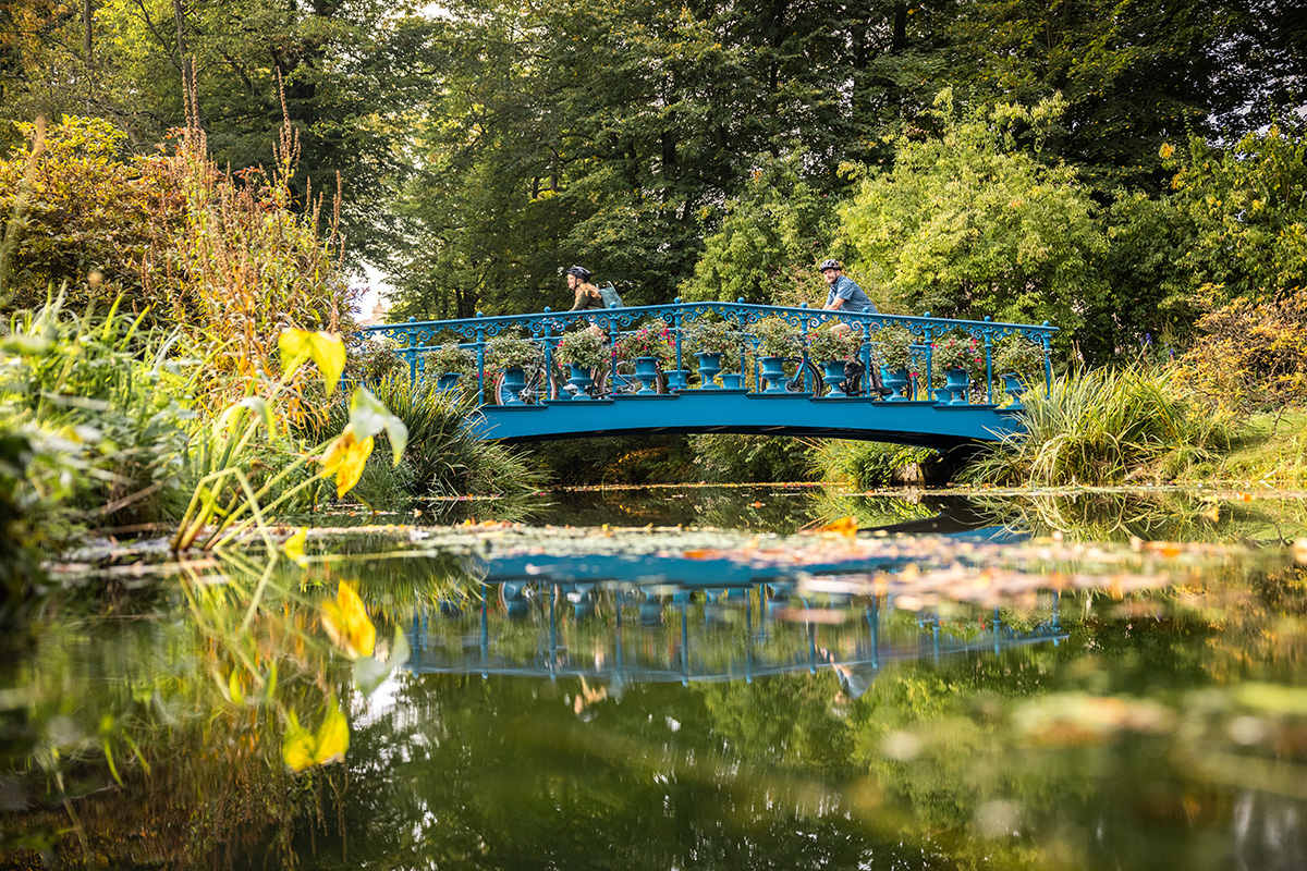 Zwei Personen fahren mit Fahrrädern über eine kleine, hellblaue Brücke mit verzierten Geländern. Auf der Brücke stehen zahlreiche Blumentöpfe mit blühenden Pflanzen. Die Brücke überspannt einen schmalen, ruhigen Fluss, in dem sich die Umgebung spiegelt. Am Ufer sind dichte, grüne Pflanzen. Im Hintergrund stehen zahlreiche Laubbäume.