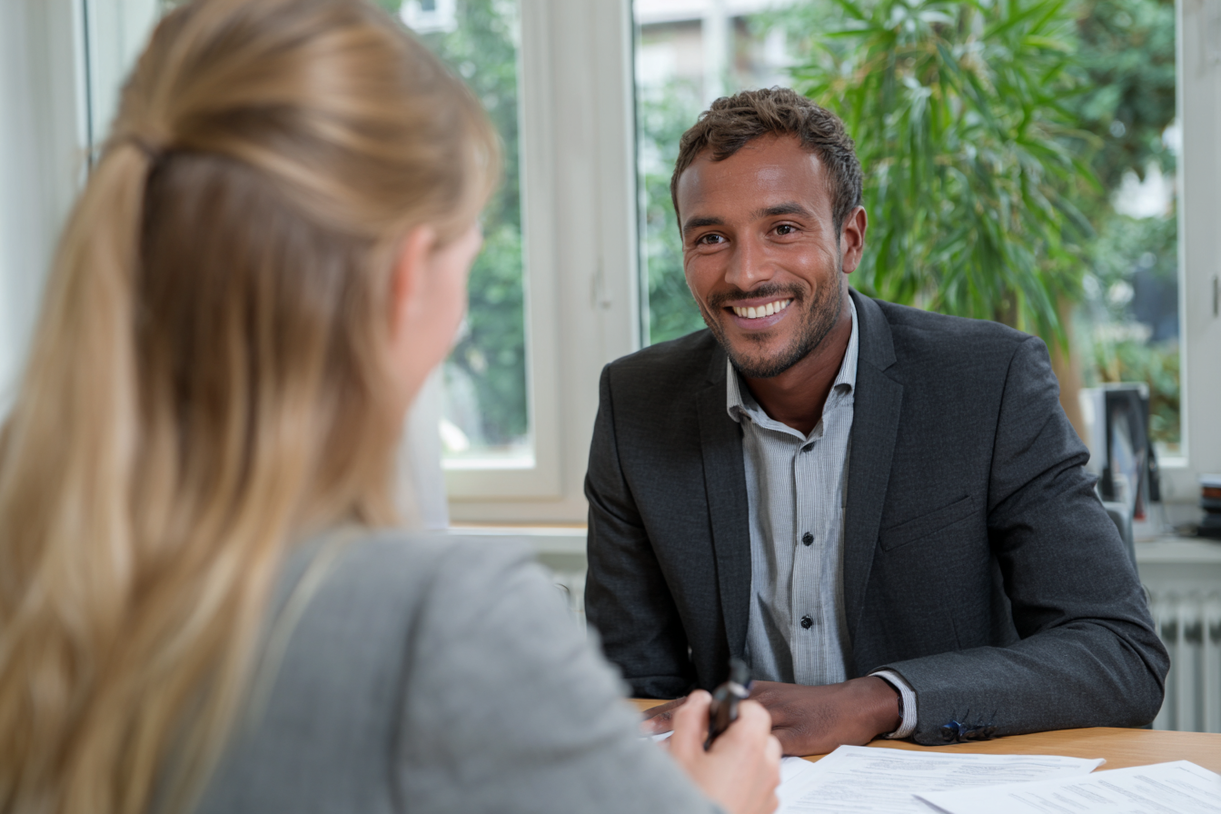 The picture shows a man sitting opposite someone in an office, smiling warmly. He appears engrossed in a consultation. The surroundings are bright and modern, with large windows and some greenery in the background.