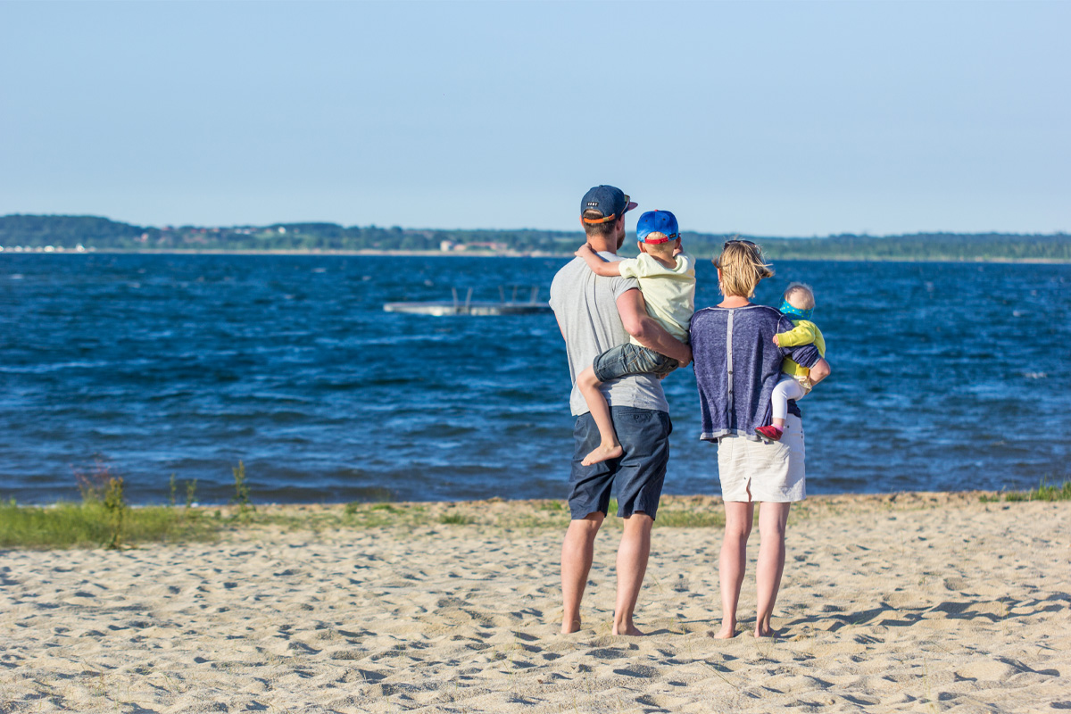 Das Bild zeigt zwei erwachsene Personen am Ufer eines Gewässers. Die beiden stehen nebeneinander auf einem Sandstrand und tragen jeweils ein Kind auf dem Arm. Alle sind von hinten zu sehen und blicken auf das Wasser. Im Hintergrund ist eine schwimmende Plattform zu erkennen, dahinter das gegenüberliegende Ufer mit bewaldeten Flächen und einzelnen Gebäuden. Der Himmel ist klar und blau.